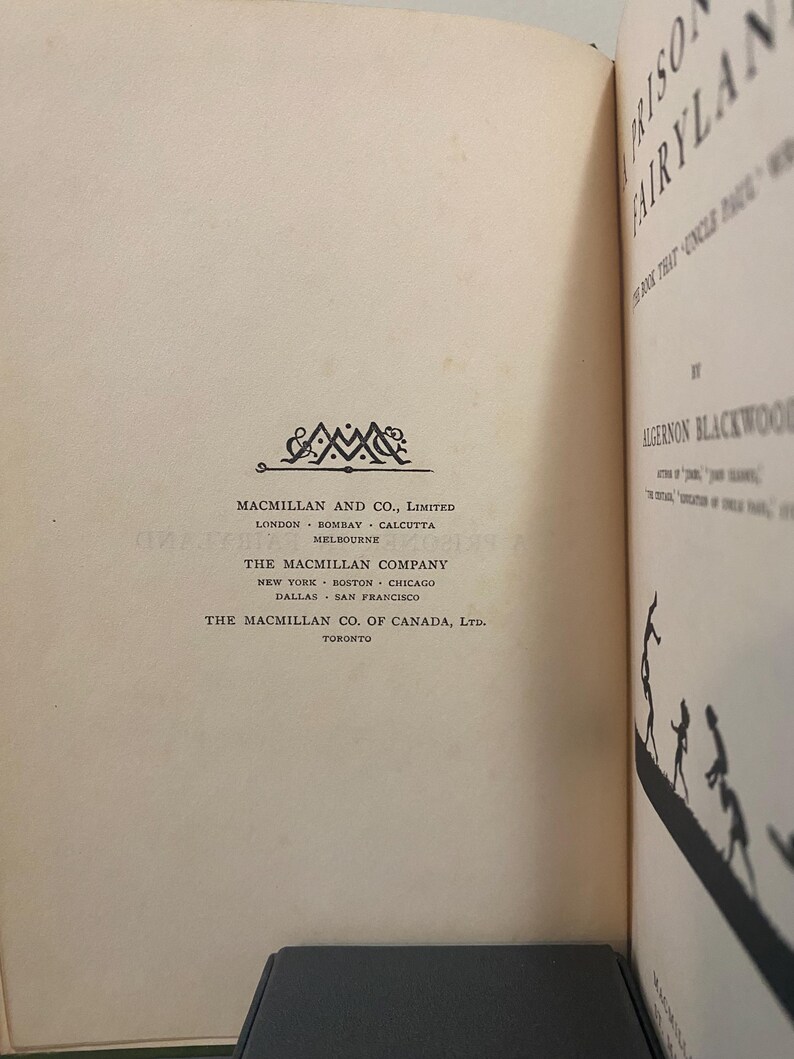 May include: Open book with the title "Prison FairyLand" by Algernon Blackwood. The book's pages are off-white with text and a decorative emblem. The Macmillan Company logo is visible.
