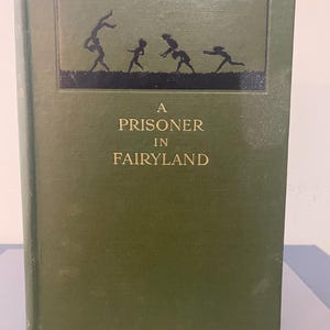May include: A hardcover book titled "A Prisoner in Fairyland" with a dark green cover. The cover features a black silhouette illustration of children playing. The title is in gold lettering.