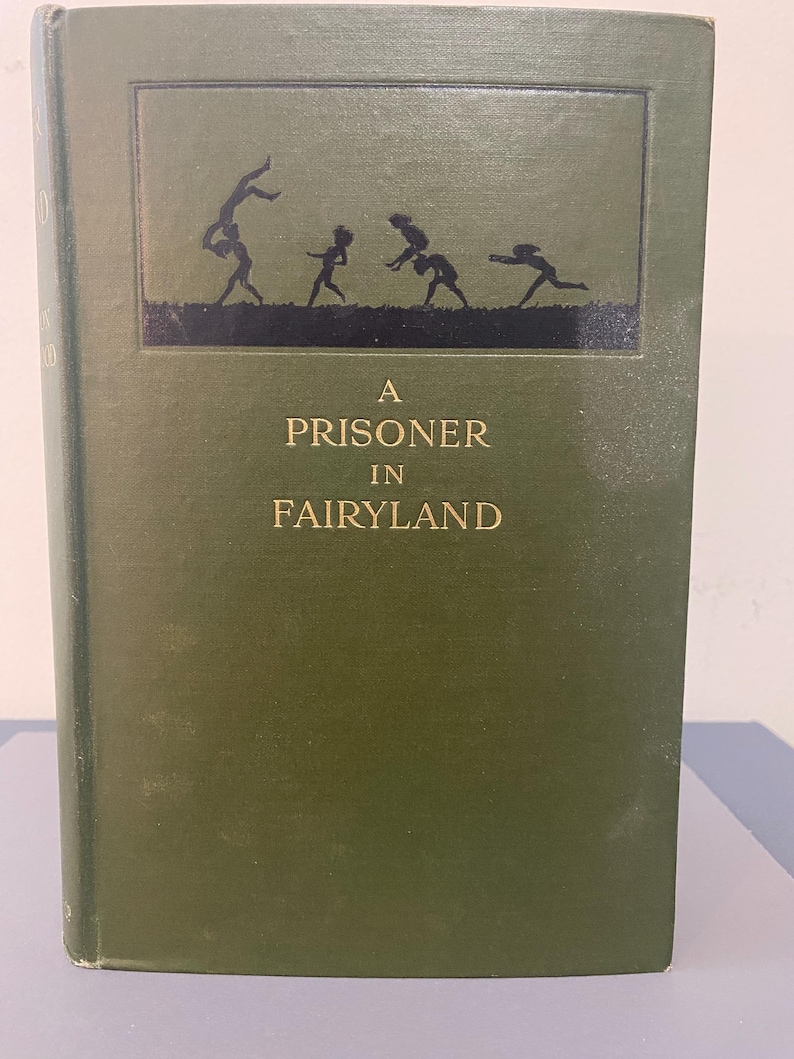 May include: A hardcover book titled "A Prisoner in Fairyland" with a dark green cover. The cover features a black silhouette illustration of children playing. The title is in gold lettering.