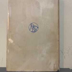 May include: A vintage book with a light beige cover, encased in clear plastic. A blue circular emblem of a person riding a horse is centered on the cover. The book's spine and edges show signs of wear.