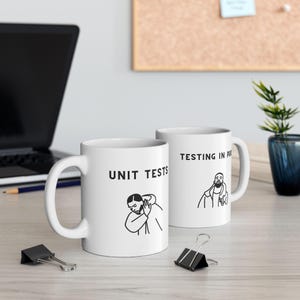 May include: Two white ceramic mugs with black text and illustrations. The mug on the left says "UNIT TESTS" and features a cartoon of a person with their head in their hands. The mug on the right says "TESTING IN PRODUCTION" and features a cartoon of a person with their hands on their hips.
