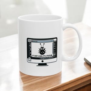 May include: White ceramic mug with a black and white illustration of a computer screen with a bug inside. The text on the screen reads "It's not a bug, it's a feature."