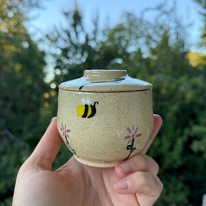 May include: A small, speckled, light yellow ceramic jar with a matching lid. The exterior features hand-painted illustrations of a black and yellow bumblebee and two purple flowers with green stems, extending from the base up to the lid.