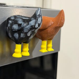 May include: Two chicken-shaped magnets. One is black with white paint, the other is brown. Both have yellow legs. They are attached to a stainless steel surface. The magnets are a fun and quirky kitchen or office decor item.