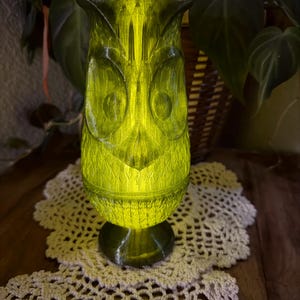 May include: A green owl-shaped lamp with a textured surface, illuminated from within. The lamp has large, round eyes and a pointed beak. It sits on a dark green base, and is placed on a cream-colored, crocheted doily.