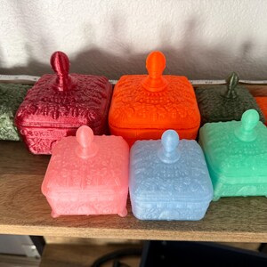 May include: A collection of square lidded boxes in various colors including red, orange, pink, blue, green, and teal. Each box features a textured, embossed surface pattern and a central knob handle on the lid, arranged in a group on a shelf.