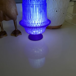 May include: A blue, illuminated lamp with a decorative, geometric design. The lamp has a flared base and a textured, cylindrical shade. The light casts a bright blue glow, creating a soft ambiance. The lamp is approximately 6 inches tall.