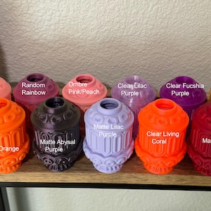 May include: A collection of colorful, decorative objects in various shades, including pink, purple, orange, and red. Each piece has a unique design and is labeled with its color name. The objects are arranged on a wooden shelf.