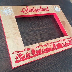 May include: Wooden picture frame with a red interior and a light wood exterior. The word "Switzerland" is in red script at the top, with a red mountain scene at the bottom. White edelweiss flowers are on the frame.