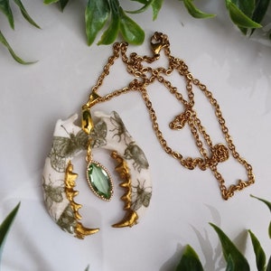 May include: A unique gold-tone necklace featuring a pendant shaped like a jawbone. The pendant is cream-colored with green insect and butterfly illustrations and gold accents. A green gemstone hangs from the center of the pendant. The necklace chain is gold.