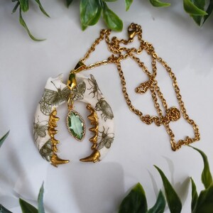 May include: Gold-toned necklace featuring a pendant shaped like a jawbone, adorned with insect illustrations. The pendant has gold accents and a green gemstone. The necklace is on a white surface with green foliage.