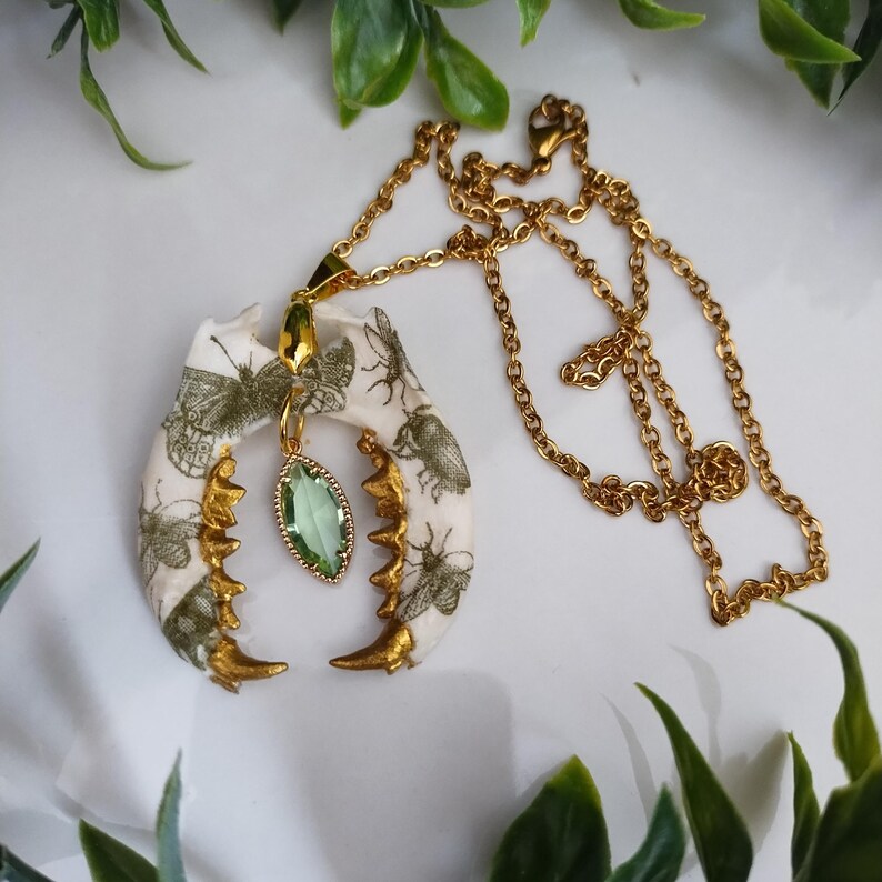 May include: Gold-toned necklace featuring a pendant shaped like a jawbone, adorned with insect illustrations. The pendant has gold accents and a green gemstone. The necklace is on a white surface with green foliage.