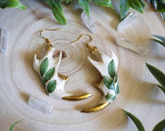 May include: A pair of unique earrings featuring a bone-like design with green leaf accents and gold-colored detailing. The earrings hang from gold-colored hooks, showcasing a nature-inspired aesthetic. The earrings are displayed on a wooden surface.