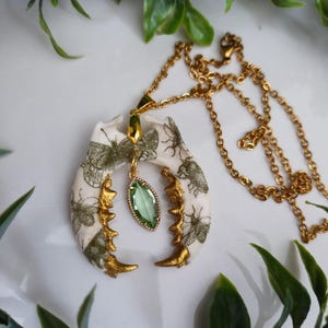 May include: A unique necklace featuring a bone-shaped pendant with insect illustrations in green and gold accents. The pendant is suspended from a gold chain and includes a green gemstone. The necklace is a statement piece.