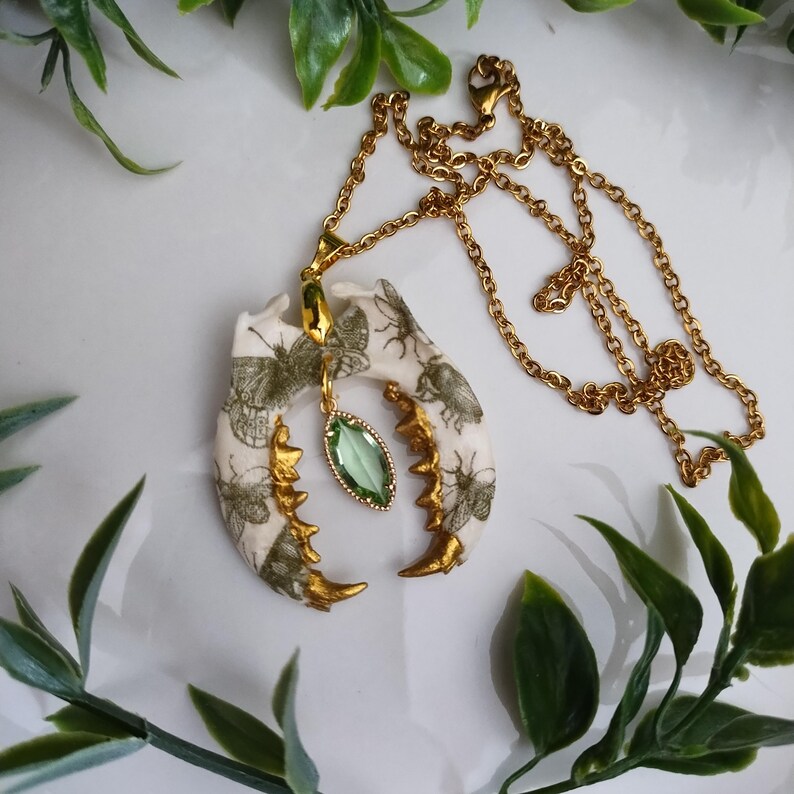 May include: A unique gold-toned necklace featuring a pendant shaped like a jawbone. The pendant is cream-colored with green insect illustrations and gold accents. A green gemstone hangs below the jawbone. The necklace chain is also gold-toned.