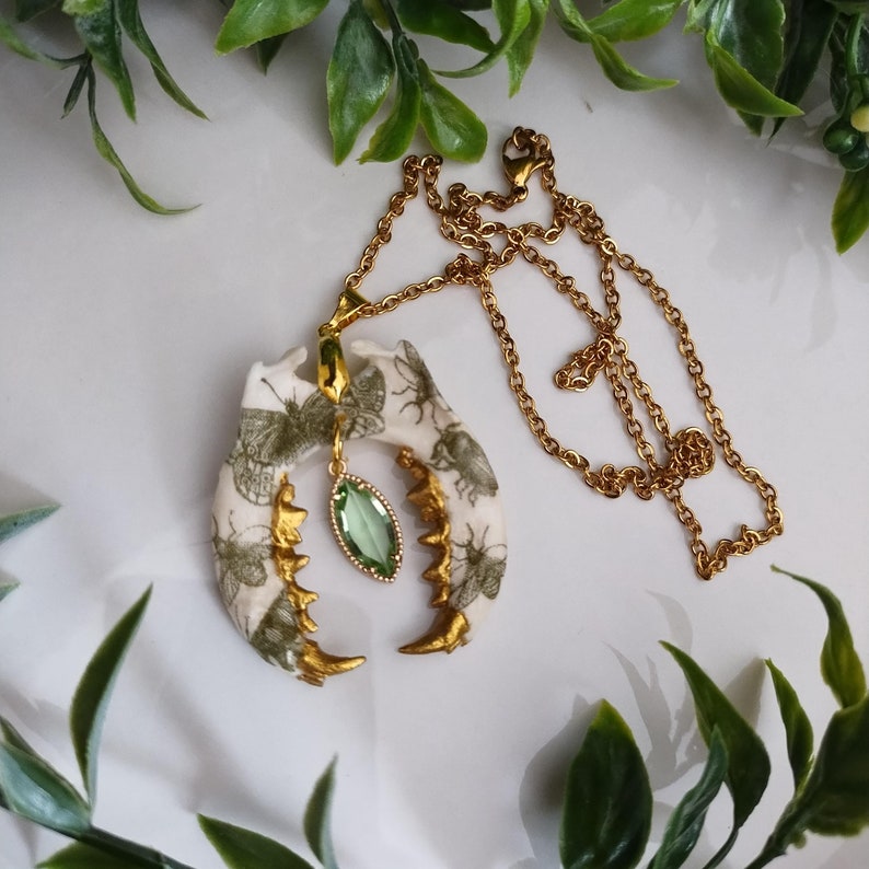 May include: A unique gold-tone necklace featuring a pendant shaped like a jawbone. The pendant is cream-colored with green insect and butterfly illustrations and gold accents. A green gemstone hangs from the center of the pendant. The necklace chain is gold.