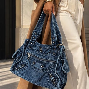 K&ouml;nnte beinhalten: Eine dunkelblaue Handtasche aus Acid-Wash-Denim mit einem silberfarbenen Metall-Logo in Form eines B, einer Rei&szlig;verschlusstasche vorne, zwei Seitentaschen mit Schnallenverschluss und zwei Schulterriemen mit silbernen Nieten.
