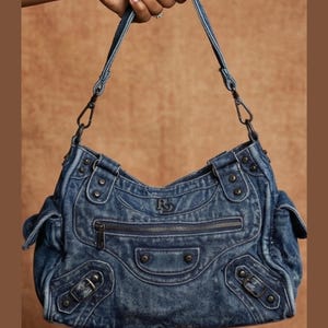 K&ouml;nnte beinhalten: Eine mittelblaue Denim-Schultertasche mit verwaschener Textur, einer Rei&szlig;verschlusstasche auf der Vorderseite und dekorativen Metallnieten. Die Tasche verf&uuml;gt &uuml;ber zwei Seitentaschen mit Klappen, verstellbare Schnallenriemen und ein Logo.