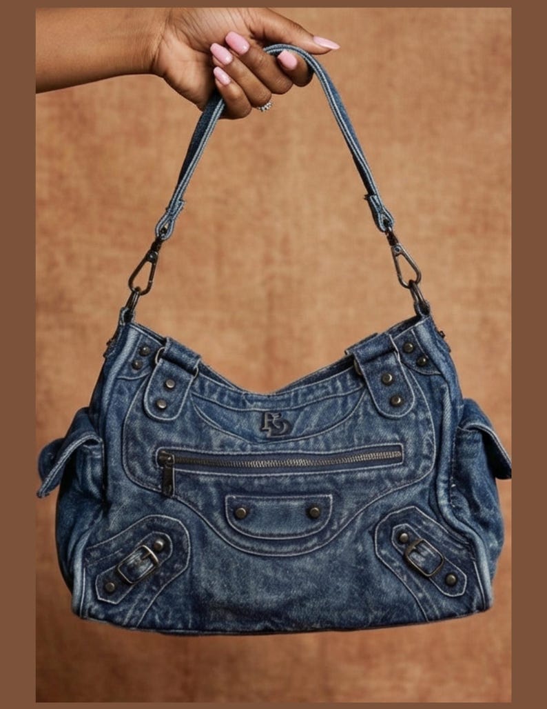 K&ouml;nnte beinhalten: Eine mittelblaue Denim-Schultertasche mit verwaschener Textur, einer Rei&szlig;verschlusstasche auf der Vorderseite und dekorativen Metallnieten. Die Tasche verf&uuml;gt &uuml;ber zwei Seitentaschen mit Klappen, verstellbare Schnallenriemen und ein Logo.