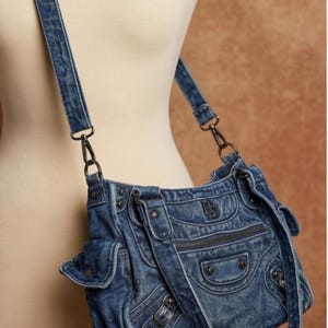 K&ouml;nnte beinhalten: Eine blaue Umh&auml;ngetasche aus Denim mit verwaschener Optik, mehreren Au&szlig;entaschen mit Klappverschl&uuml;ssen, einem vorderen Rei&szlig;verschlussfach, dunklen Metallbeschl&auml;gen und einem verstellbaren Denim-Schulterriemen mit Karabinerhaken aus Metall.