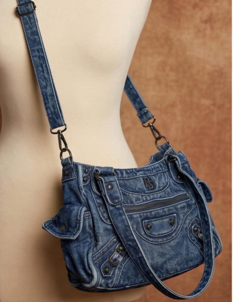 K&ouml;nnte beinhalten: Eine blaue Umh&auml;ngetasche aus Denim mit verwaschener Optik, mehreren Au&szlig;entaschen mit Klappverschl&uuml;ssen, einem vorderen Rei&szlig;verschlussfach, dunklen Metallbeschl&auml;gen und einem verstellbaren Denim-Schulterriemen mit Karabinerhaken aus Metall.
