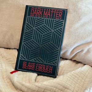 May include: A hardcover book titled "DARK MATTER" by Blake Crouch. The book has a dark teal cover with silver geometric patterns and red lettering. A red ribbon bookmark is visible.