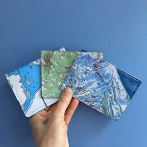 May include: Three square coasters with map designs. One coaster features a blue lake map, another a green topographic map, and the third a ski resort map with blue and white slopes. Each coaster is stitched with white thread.