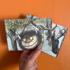May include: Two Where the Wild Things Are themed wallets. The wallets feature a monster illustration with a black, white, and yellow color scheme. The wallets are held against an orange background.