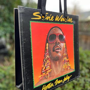 May include: Black tote bag featuring the album art for Stevie Wonder's "Hotter Than July." The artwork shows a portrait of Stevie Wonder with red sunglasses against a red and orange background. The bag has black handles.