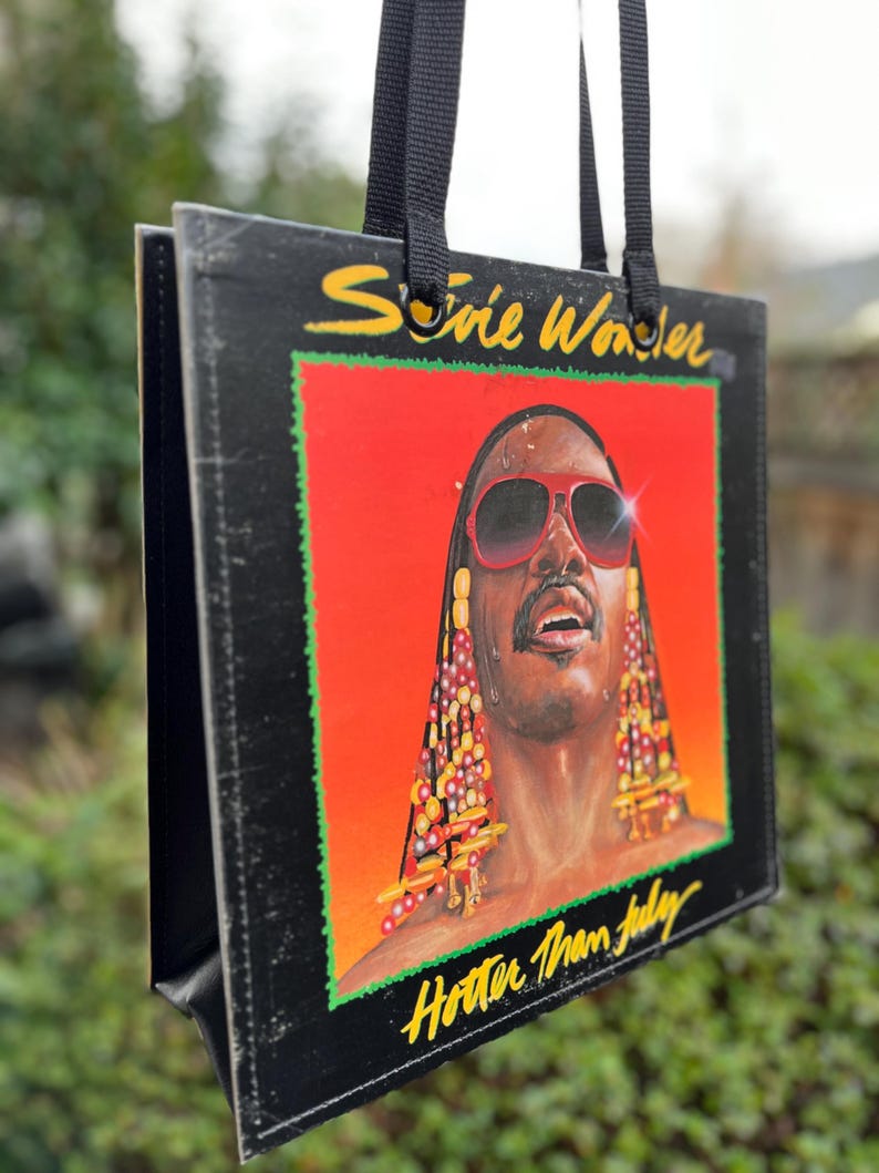 May include: Black tote bag featuring the album art for Stevie Wonder's "Hotter Than July." The artwork shows a portrait of Stevie Wonder with red sunglasses against a red and orange background. The bag has black handles.