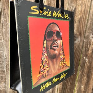 May include: Black tote bag featuring the album art from Stevie Wonder's "Hotter Than July." The artwork shows a portrait of Stevie Wonder wearing sunglasses against a red and orange background. The bag has black handles and a black border.