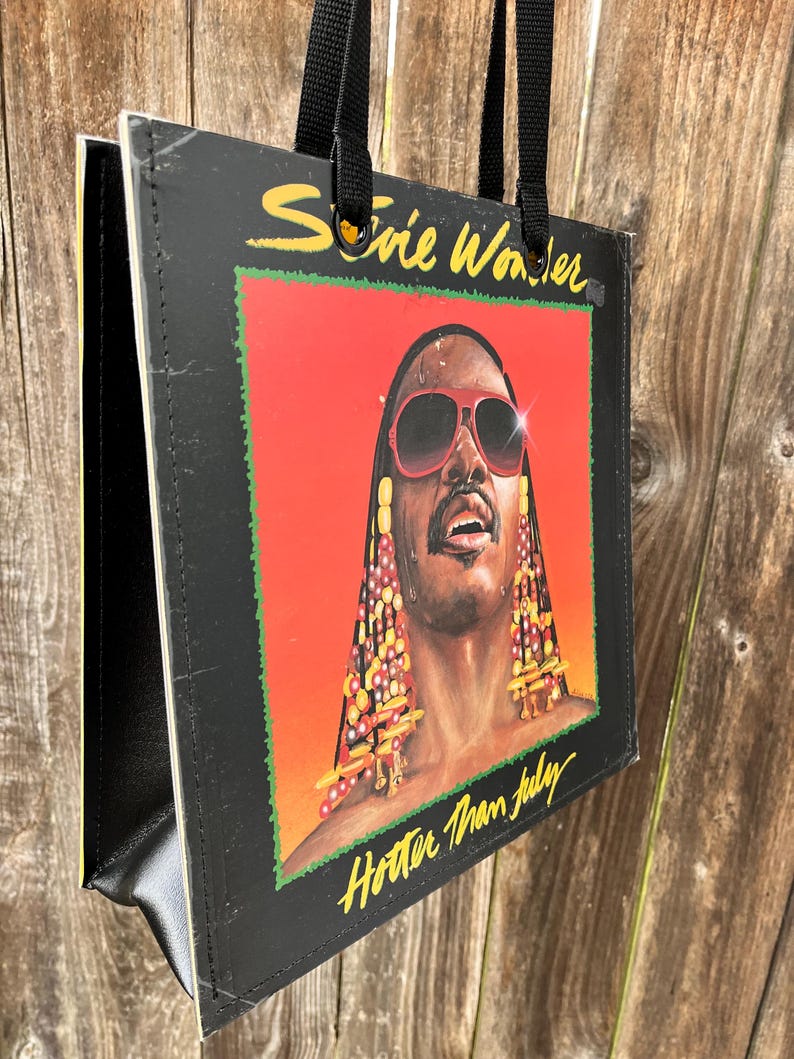 May include: Black tote bag featuring the album art from Stevie Wonder's "Hotter Than July." The artwork shows a portrait of Stevie Wonder wearing sunglasses against a red and orange background. The bag has black handles and a black border.