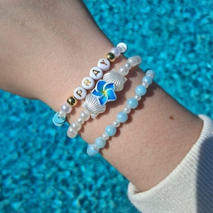 May include: Three beaded bracelets. One bracelet spells "PRAY" with white letter beads and gold accents. Another features white pearl beads, seashell charms, and a blue flower. The third bracelet has light blue beads.