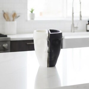 May include: A decorative ceramic vase with a face design, split vertically into black and white halves. The vase is displayed on a white countertop in a modern kitchen setting. Green apples and a small succulent are also visible.