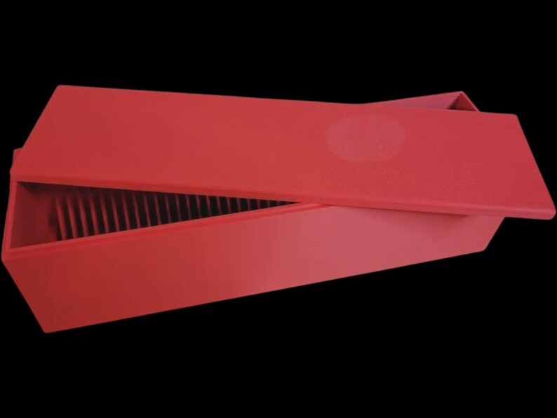 Puede incluir: Una caja rectangular roja con una tapa ligeramente abierta, que revela divisores verticales en el interior. La caja y la tapa son de un rojo mate s&oacute;lido sobre un fondo negro. La tapa tiene una hendidura circular.