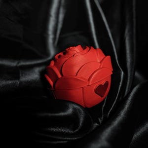 May include: A red, rose-shaped box with a heart cutout on the side, set against a backdrop of black satin fabric. The box is a vibrant red, and the satin creates a dramatic contrast.