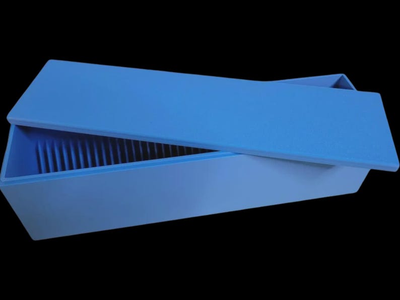 Puede incluir: Una caja rectangular azul claro con una tapa a juego, parcialmente abierta. El interior de la caja tiene crestas verticales. La caja est&aacute; sobre un fondo negro.
