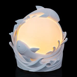 May include: White decorative lamp featuring two fish leaping over a glowing orb, surrounded by stylized waves. The lamp is made of a solid white material and has a smooth finish. The overall design evokes a sense of aquatic life and tranquility.