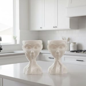 May include: Two white ceramic face planters with closed eyes and hands supporting the face. The planters are on a white countertop in a kitchen setting. The planters are a decorative item.