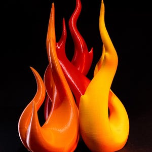 May include: A cluster of flame-shaped sculptures in vibrant colors. The forms are primarily orange, red, and yellow, with smooth, curved surfaces. The background is a solid black, highlighting the dynamic shapes and hues of the artwork.