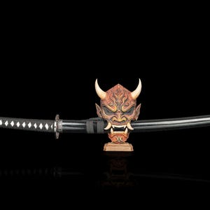 May include: A black katana sword with a wrapped handle rests horizontally on a detailed, brown-toned Hannya mask display stand featuring prominent horns, sharp teeth, and intricate facial carvings against a solid black background.