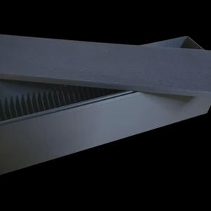 Puede incluir: Una caja rectangular gris oscuro con una tapa ligeramente entreabierta, que revela una serie de divisores verticales en el interior. La tapa es plana y texturizada, y la caja parece estar hecha de un material mate. El fondo es negro.