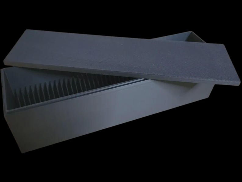 Puede incluir: Una caja rectangular gris oscuro con una tapa ligeramente entreabierta, que revela una serie de divisores verticales en el interior. La tapa es plana y texturizada, y la caja parece estar hecha de un material mate. El fondo es negro.