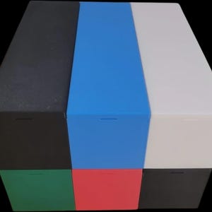 Puede incluir: Un cubo de almacenamiento multicolor con seis lados, cada uno de un color diferente: negro, azul, blanco, verde, rojo y un segundo negro. Cada lado tiene una peque&ntilde;a ranura horizontal. El cubo est&aacute; sobre un fondo negro.