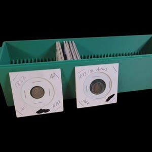 May include: A green plastic coin storage box with a lid, holding several coin holders. Two coin holders are visible, each containing a coin and handwritten details. One coin holder is labeled "1853" and the other "1973 10c Arrows".