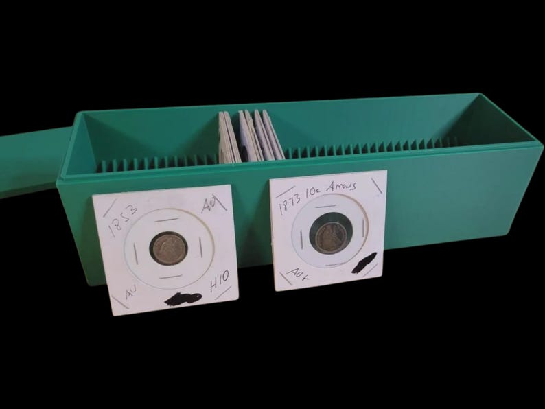 Puede incluir: Una caja verde de pl&aacute;stico para guardar monedas con tapa, que contiene varios portamonedas. Se ven dos portamonedas, cada uno con una moneda y detalles escritos a mano. Uno est&aacute; etiquetado "1853" y el otro "1973 10c Arrows".