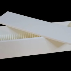 Puede incluir: Una caja rectangular blanca con una tapa ligeramente entreabierta, que revela divisores verticales en el interior. La caja y la tapa son de un blanco mate s&oacute;lido, sobre un fondo negro. El dise&ntilde;o sugiere almacenamiento.