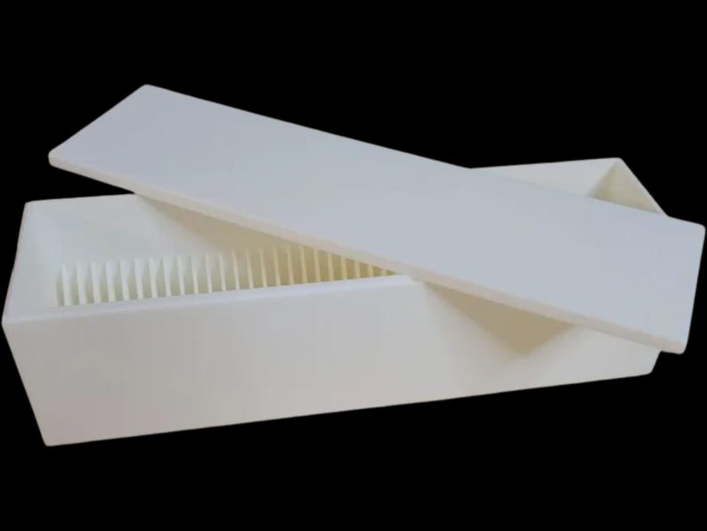 Puede incluir: Una caja rectangular blanca con una tapa ligeramente entreabierta, que revela divisores verticales en el interior. La caja y la tapa son de un blanco mate s&oacute;lido, sobre un fondo negro. El dise&ntilde;o sugiere almacenamiento.