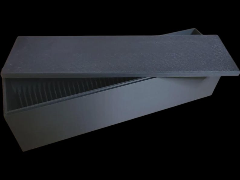 Puede incluir: Una caja rectangular gris oscuro con una tapa texturizada, parcialmente abierta para revelar el interior. La caja tiene un dise&ntilde;o inclinado y el interior tiene crestas verticales. El fondo es negro.