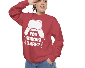 Może przedstawiać: Czerwona bluza z białym nadrukiem czapki zimowej i napisem "YOU SERIOUS CLARK?". Bluza jest w jednolitym czerwonym kolorze i ma długie rękawy. Grafika jest umieszczona na środku.